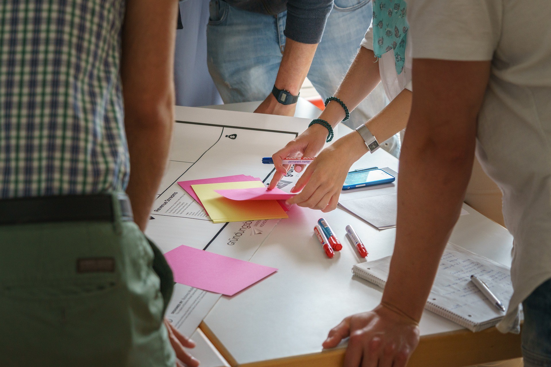Viele Hände stützen sich auf einen Tisch. Auf dem Tisch liegt Papier und zahlreiche Post-its und Stifte
