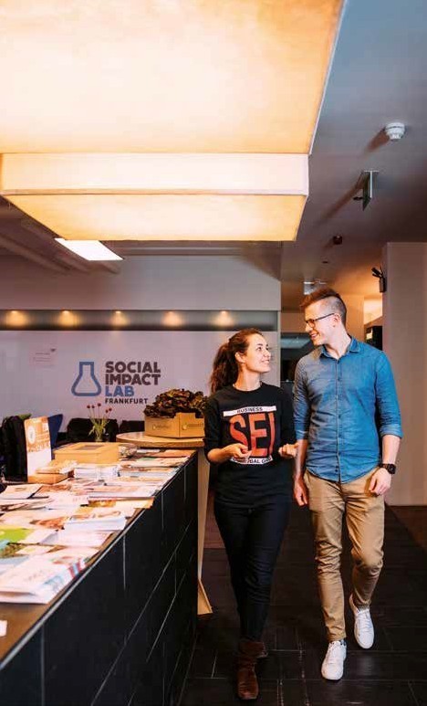 Ein junger Mann und eine junge Frau unterhalten sich im Gehen in einem gro&szlig;en B&uuml;roraum. Im Hintergrund das Schild: Social Impact Lab