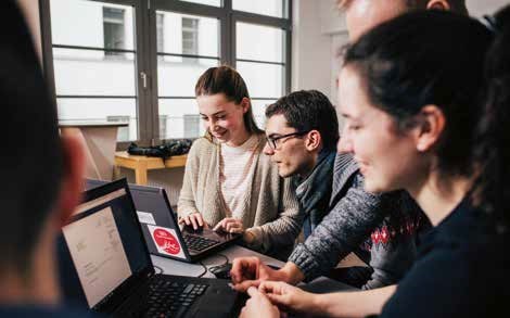 Mehrere junge Leute stehen um zwei Laptops herum. Alle sind am L&auml;cheln.
